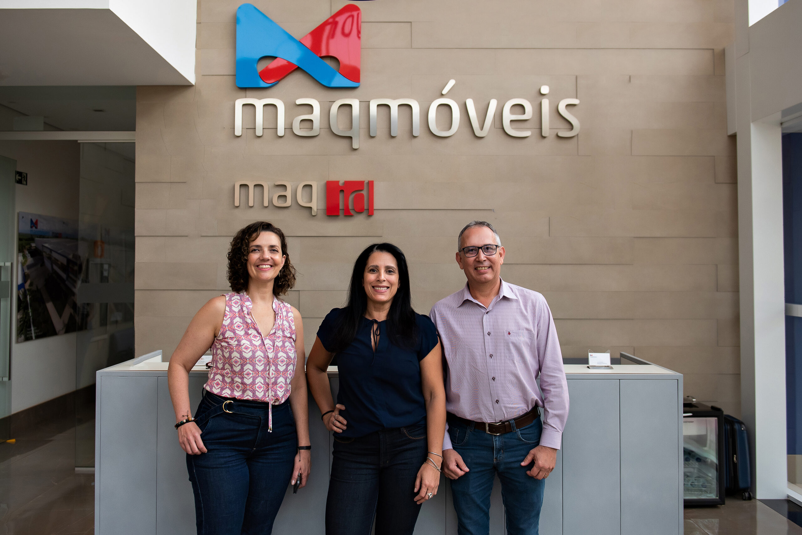 Duas mulheres e um homem sorrindo para a foto e em frente a um balcão. Na parede ao fundo, a logo da Maqmóveis.