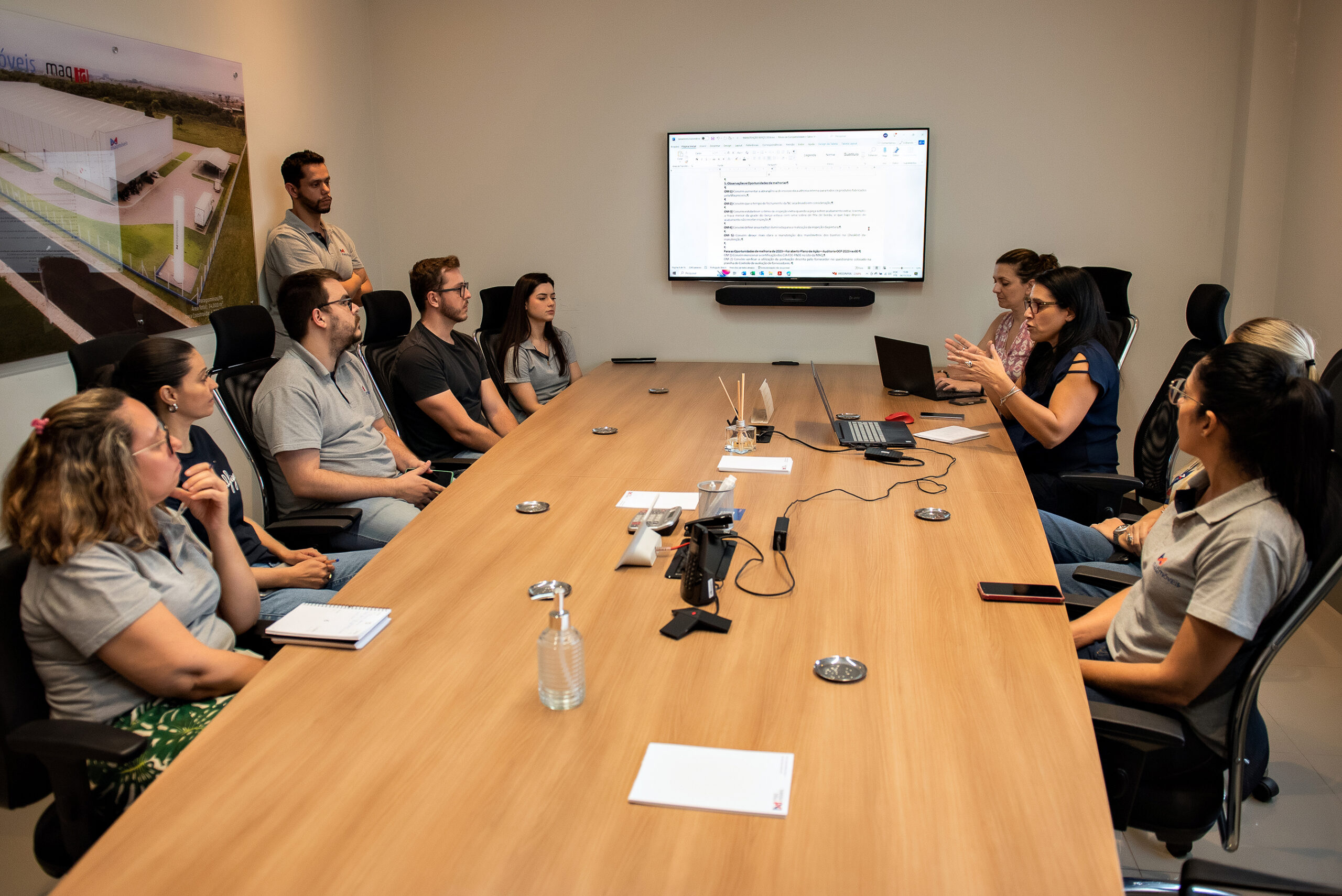 Uma mesa de reunião com notebooks e carregadores e pessoas ao redor. Ao fundo, uma televisão projeta um documento.